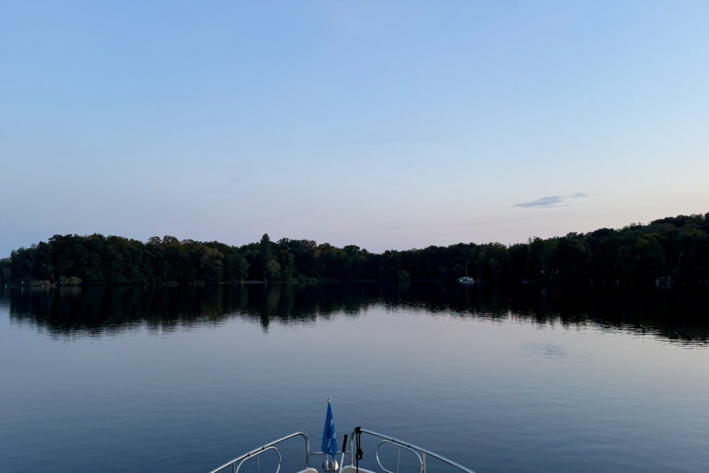 Spitze eines Bootes mit einer blauen Fahne auf einem glatten windstillen See. Im Hintergrund eine Baumreihe, die sich im Wasser spiegelt.